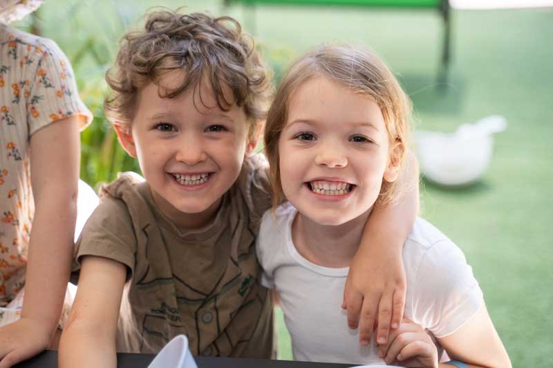 Two children grinning as they sit next to each other building social skills and finding friends. The child on the left in a brown shirt has his arm draped over the child next to him in a white shirt. Another child, partially seen stands on their left.
